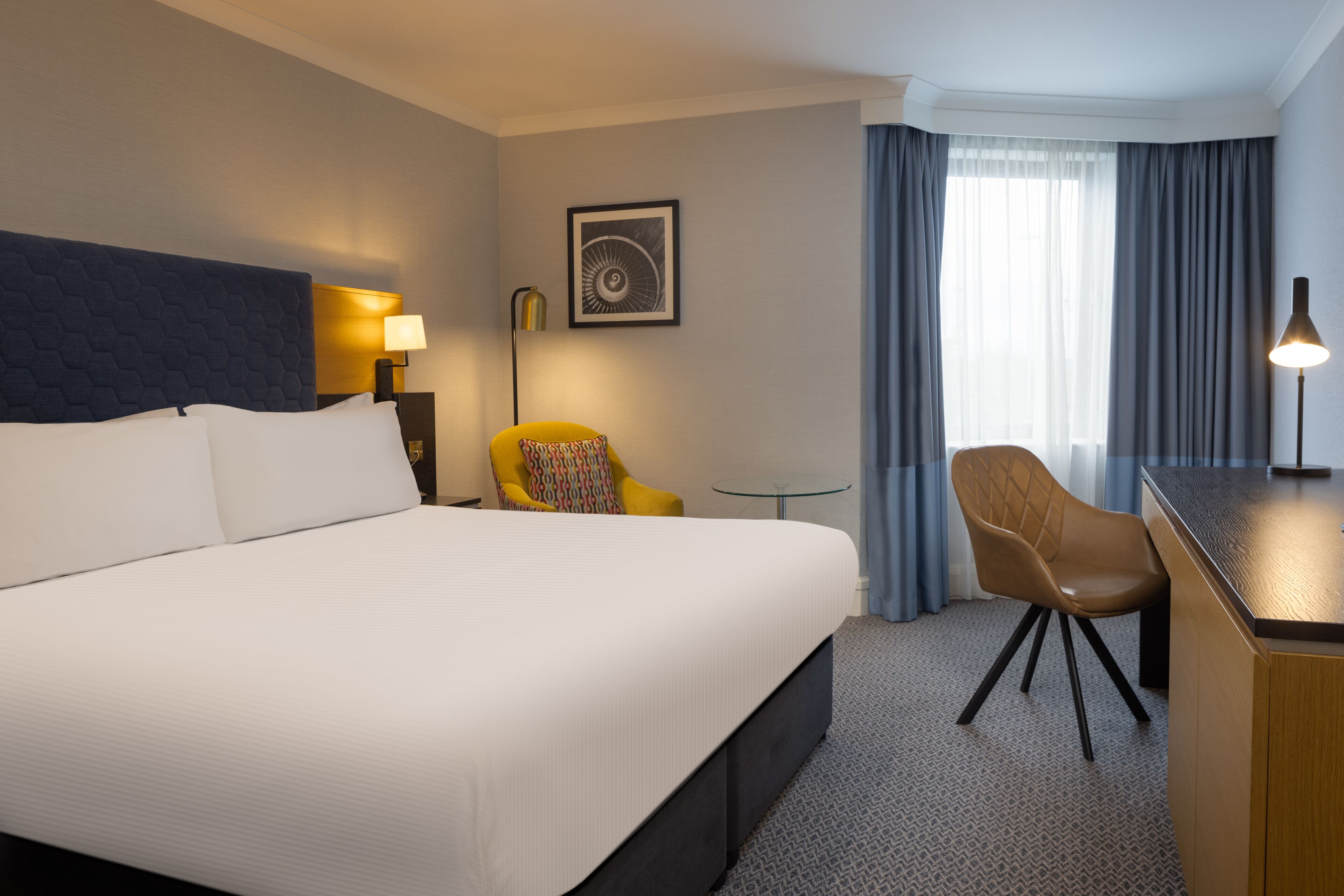 A hotel room with a large bed, a desk, a chair, a lamp, and a window with curtains.