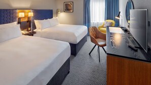 Ein Hotelzimmer mit zwei Betten, einem Schreibtisch mit Fernseher, einem Sessel und einem Fenster mit Vorhängen.