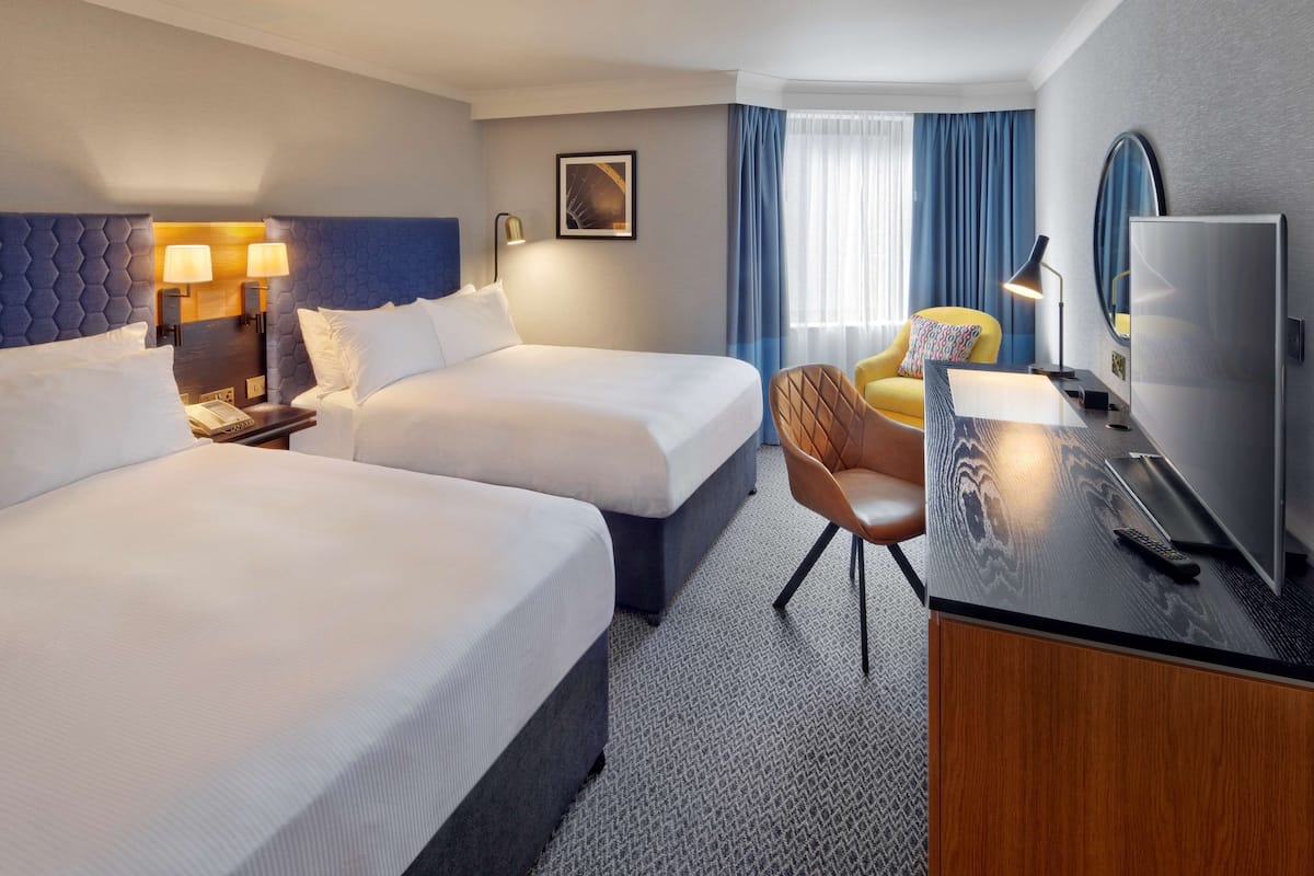 A hotel room with two beds, a desk with a TV, a chair, and a window with curtains.