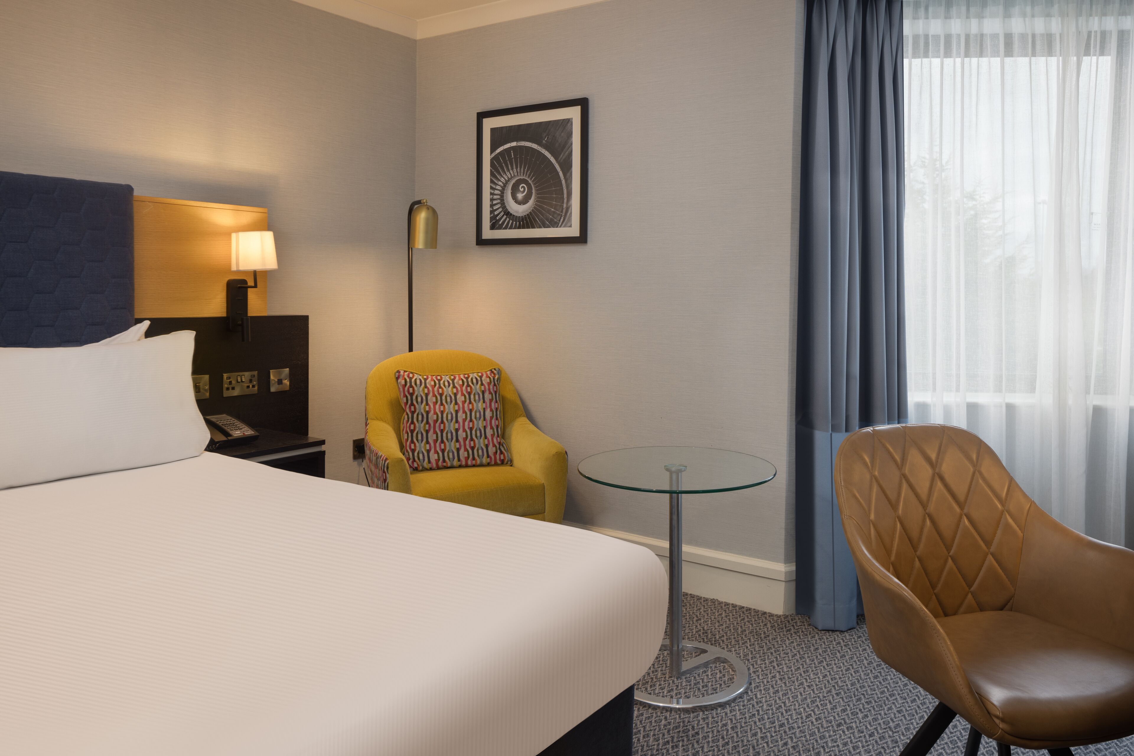 A hotel room with a bed, a desk with a lamp, a chair, a small table, and a window with curtains.