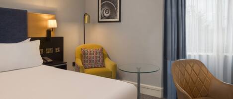 A hotel room with a bed, a desk with a lamp, a chair, a small table, and a window with curtains.