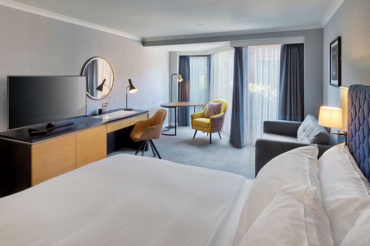 A hotel room with a large bed, a desk with a chair, a small round table, a yellow armchair, and a sofa.