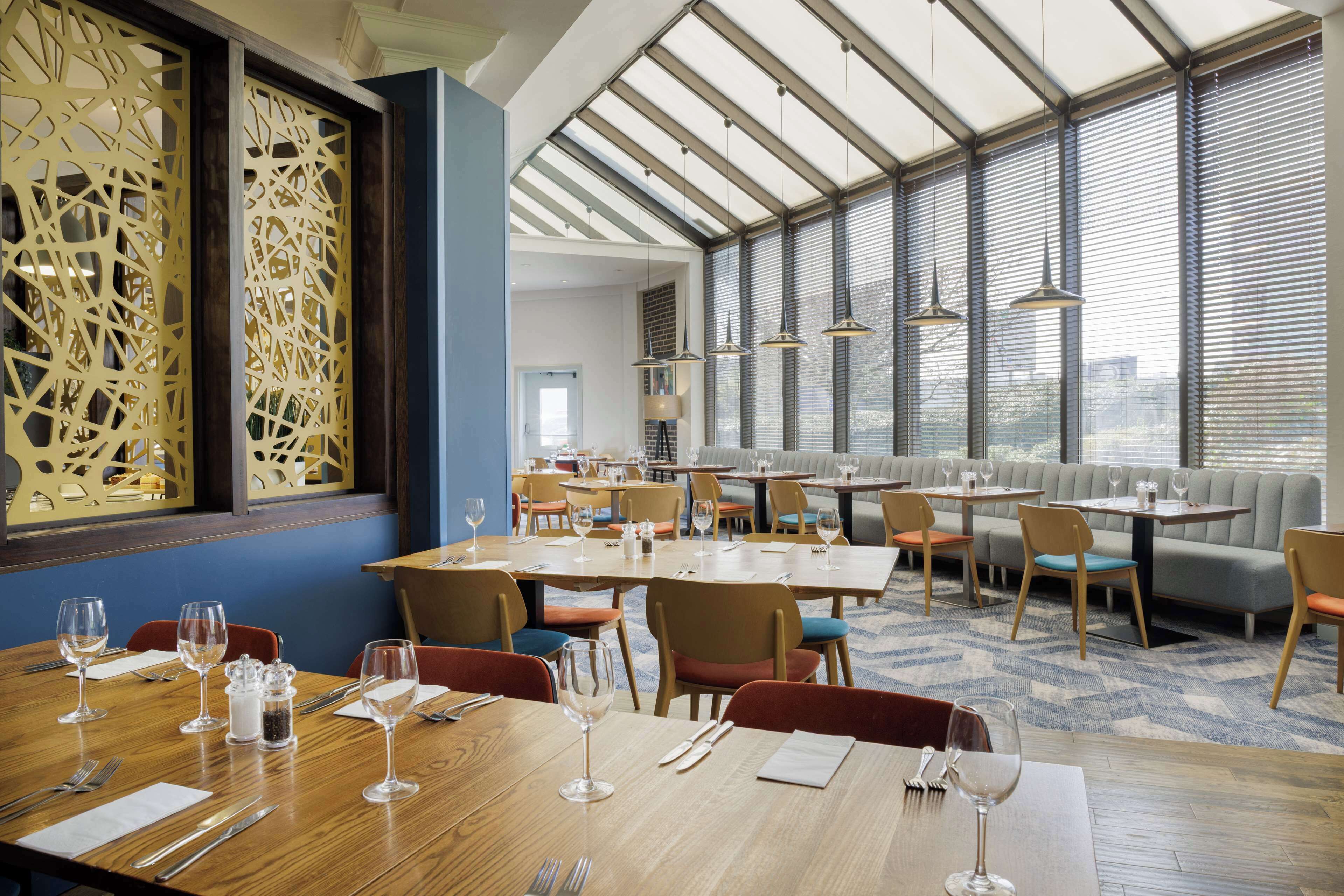 A restaurant with multiple dining tables and chairs, a large decorative screen, and large windows with blinds.