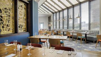 A restaurant with multiple dining tables and chairs, a large decorative screen, and large windows with blinds.