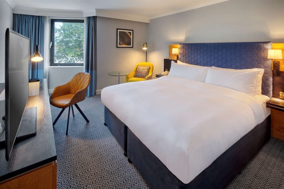 A hotel room with a large bed, a desk with a flat-screen TV, a chair, and a window with curtains.