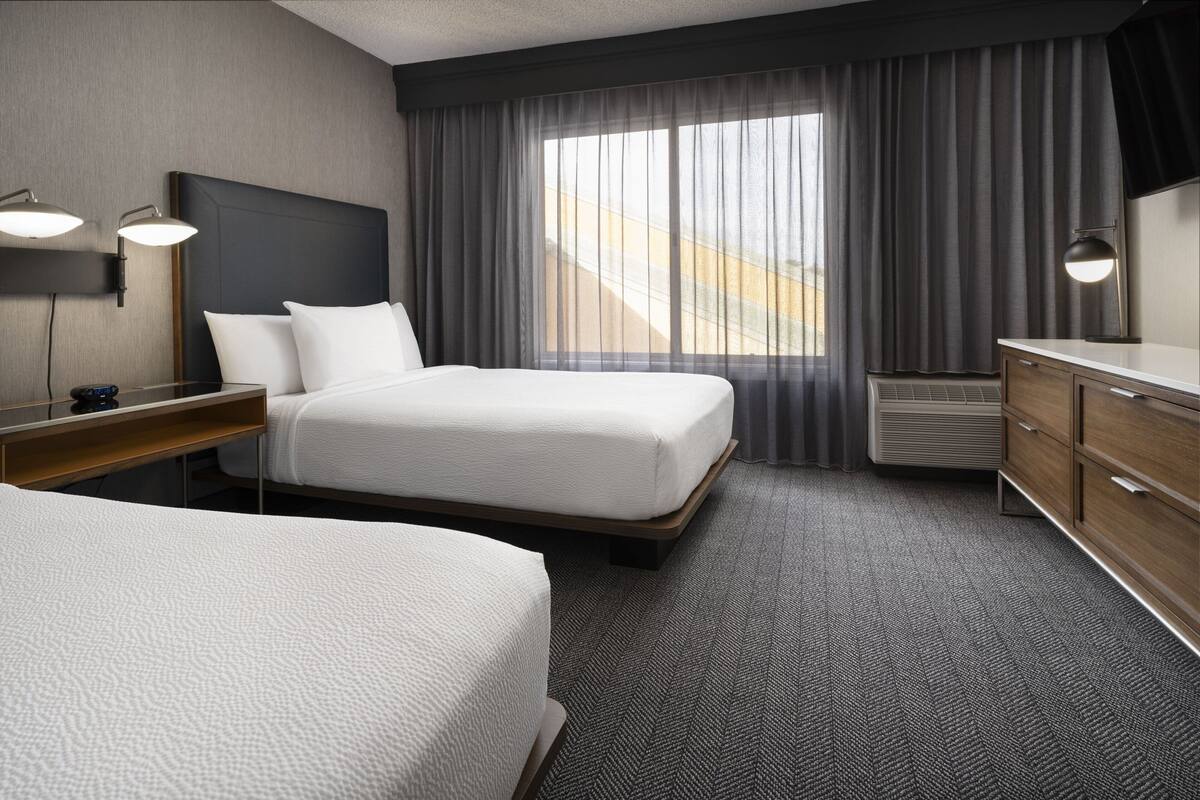 Habitación de hotel con una cama grande, otra más pequeña, un escritorio con lámpara, cómoda y un ventanal con cortinas.