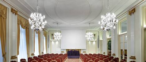 A conference room with rows of red chairs, a stage with a blue backdrop, and chandeliers.