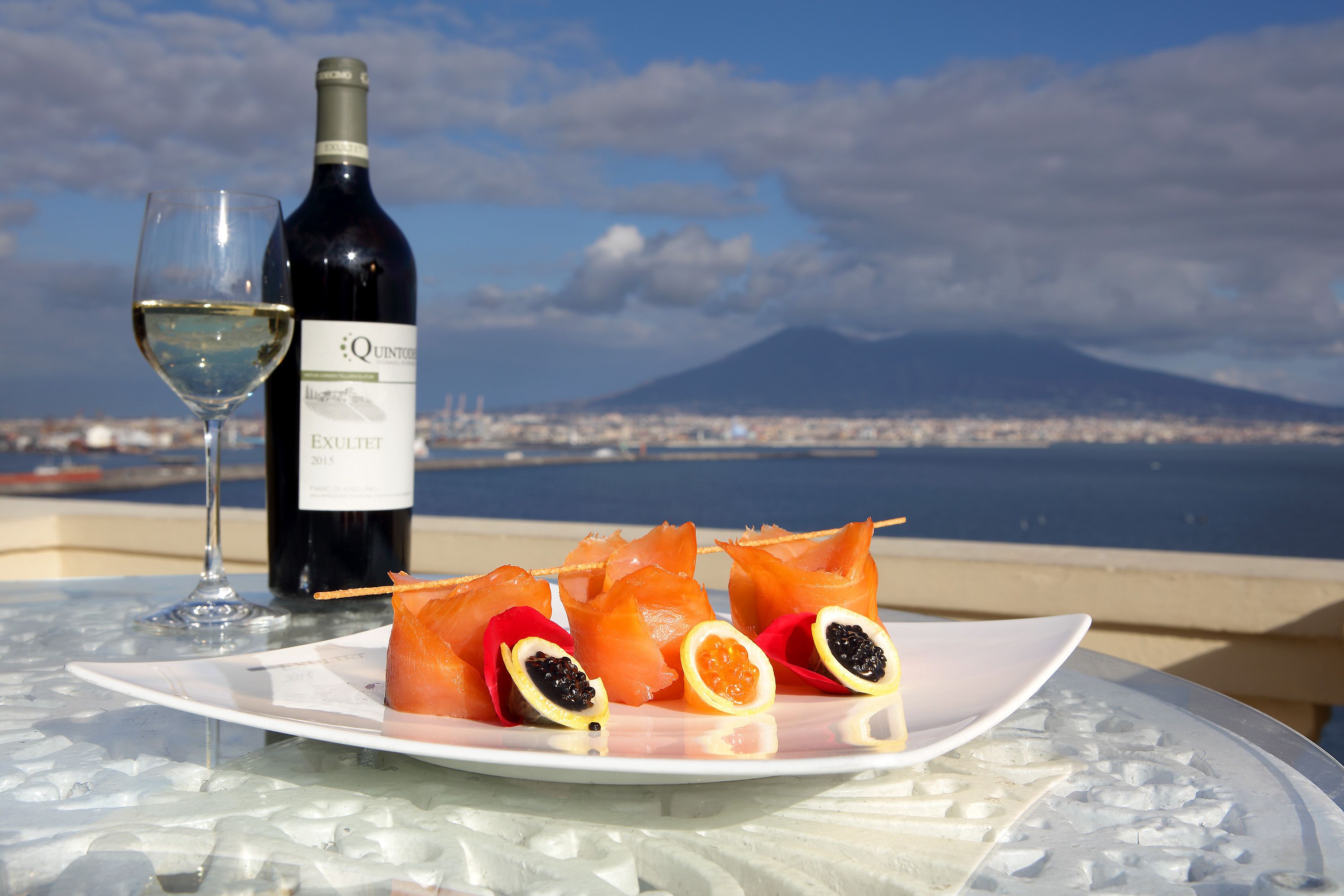 Una copa de vino blanco, una botella de vino y un plato con salmón y cítricos sobre una mesa con superficie de vidrio, con vista a una ciudad y una montaña.