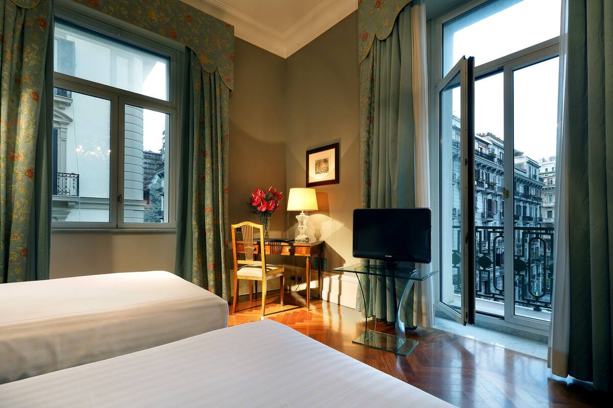 A hotel room with two beds, a desk with a chair, a TV, and a large window with a view of buildings.