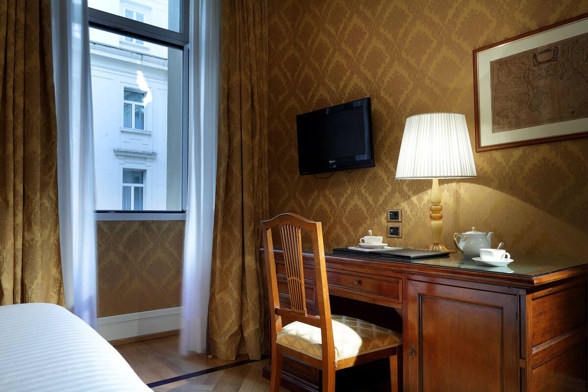 A hotel room with a bed, a desk with a lamp, a chair, a television, and a window with curtains.