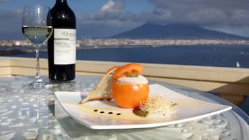Una copa de vino blanco, una botella de vino y un plato con comida sobre una mesa con superficie de vidrio, con vistas a una ciudad y una montaña.