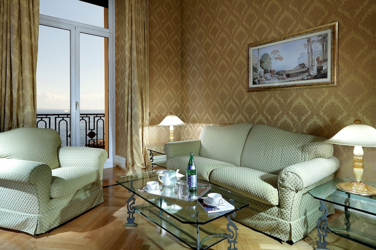 A room with a sofa, armchairs, a glass coffee table, and a painting on the wall.