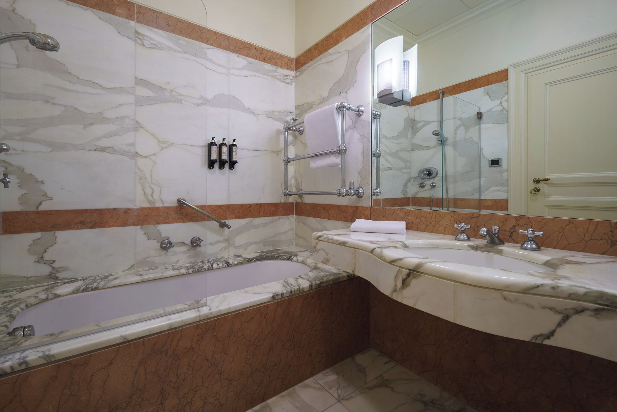 A bathroom with a marble wall, a bathtub, a shower, and a vanity with two sinks.