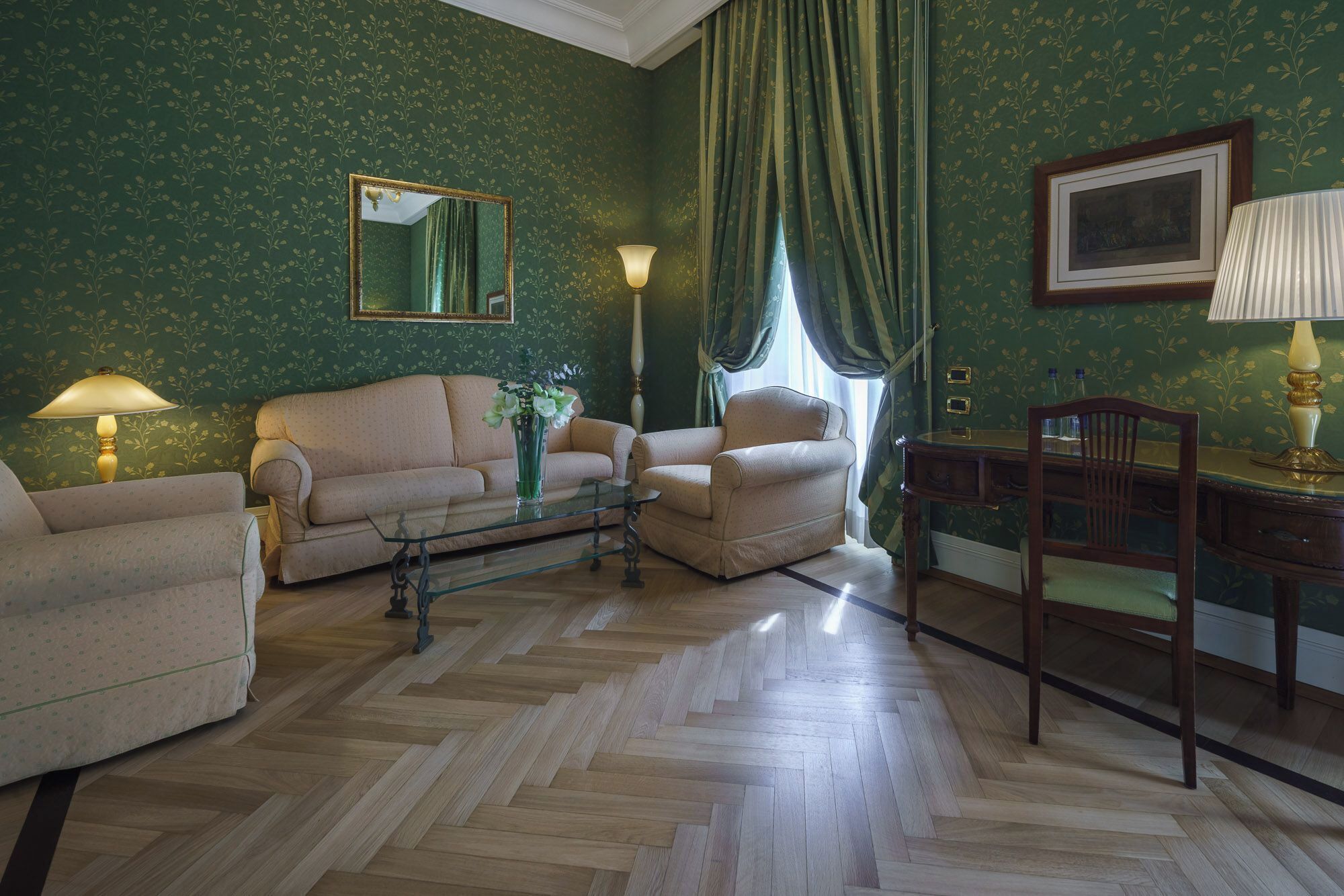 A room with a herringbone parquet floor, a sofa, a chair, a small table, and a desk with a chair.