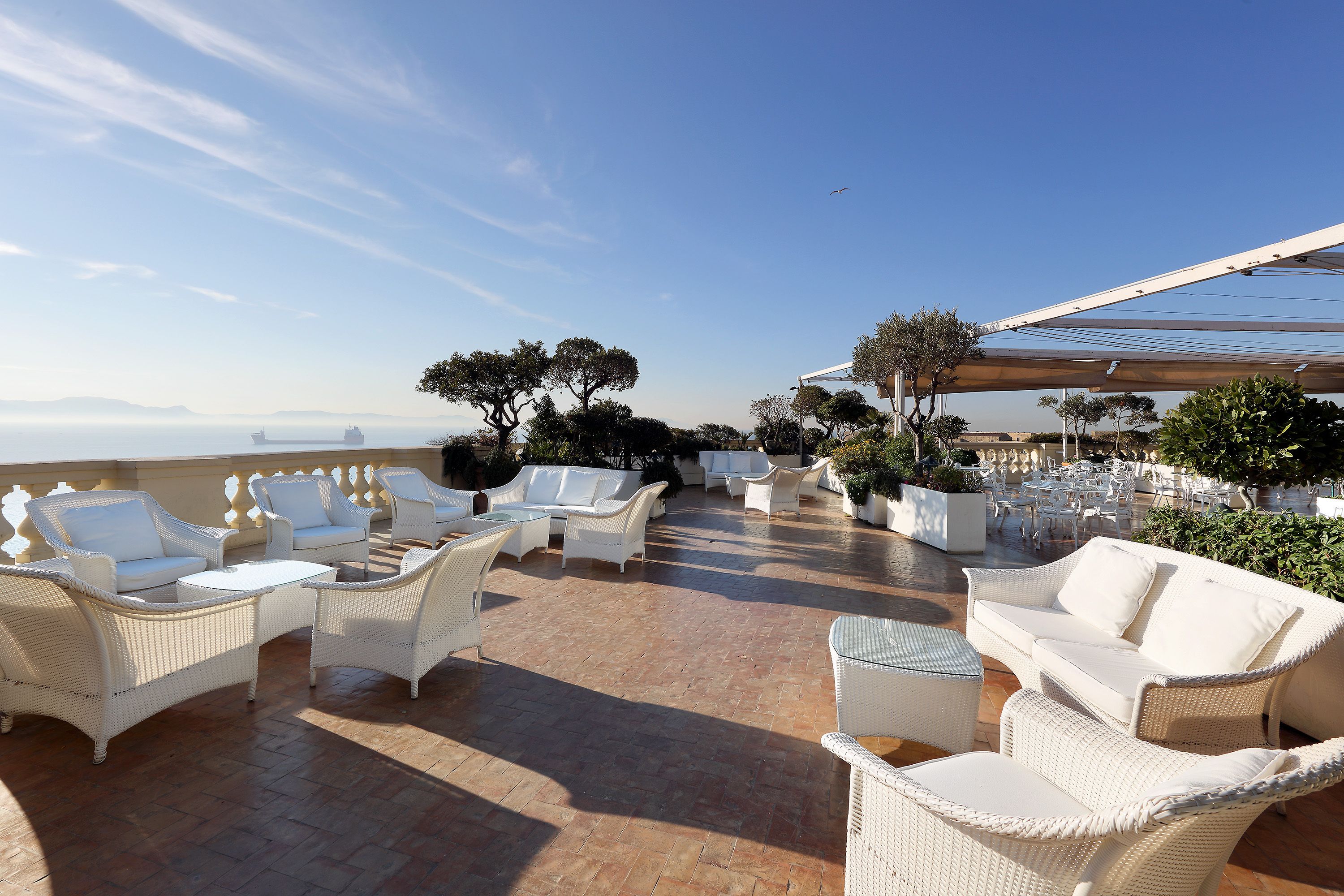 Een terras buiten met witte rieten meubels, potplanten en uitzicht op zee.
