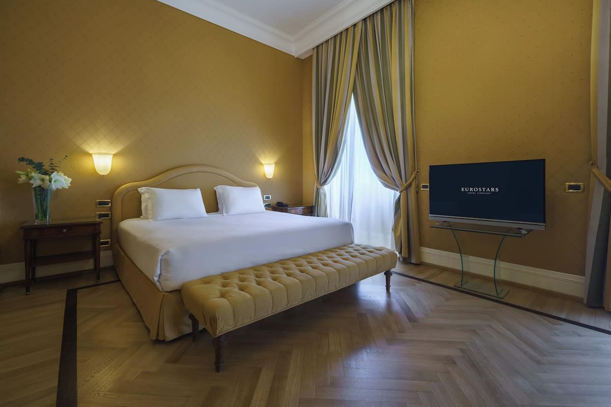 A hotel room with a large bed, a bedside table with a vase of flowers, a television on a stand, and a view of the outdoors through the window.