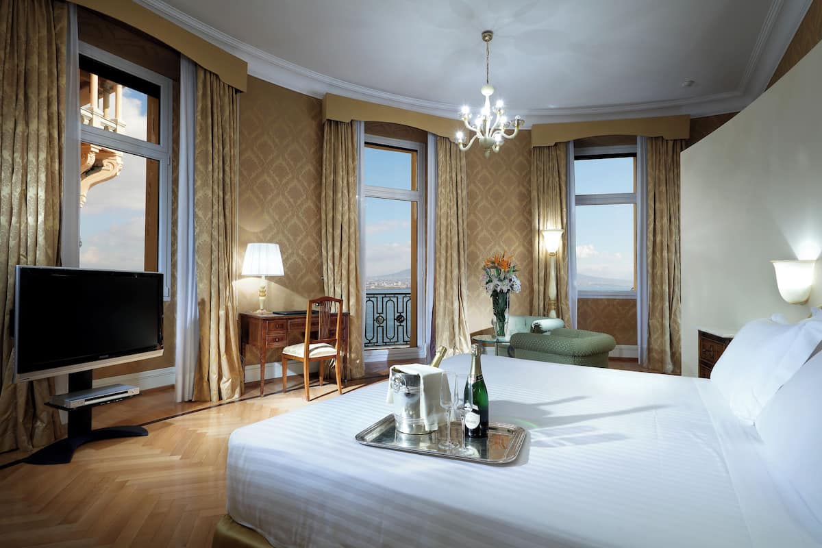 A hotel room with a large bed, a TV, a desk with a chair, and a view of the outdoors through large windows.