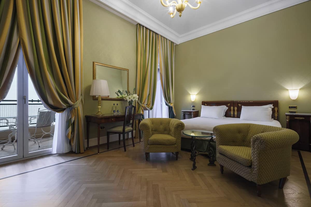 A hotel room with a bed, two armchairs, a desk with a lamp, a chair, a small table, and a large window with curtains.