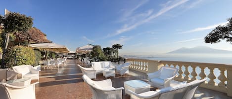 A terrace with white wicker furniture, a view of the sea, and a mountain in the background.