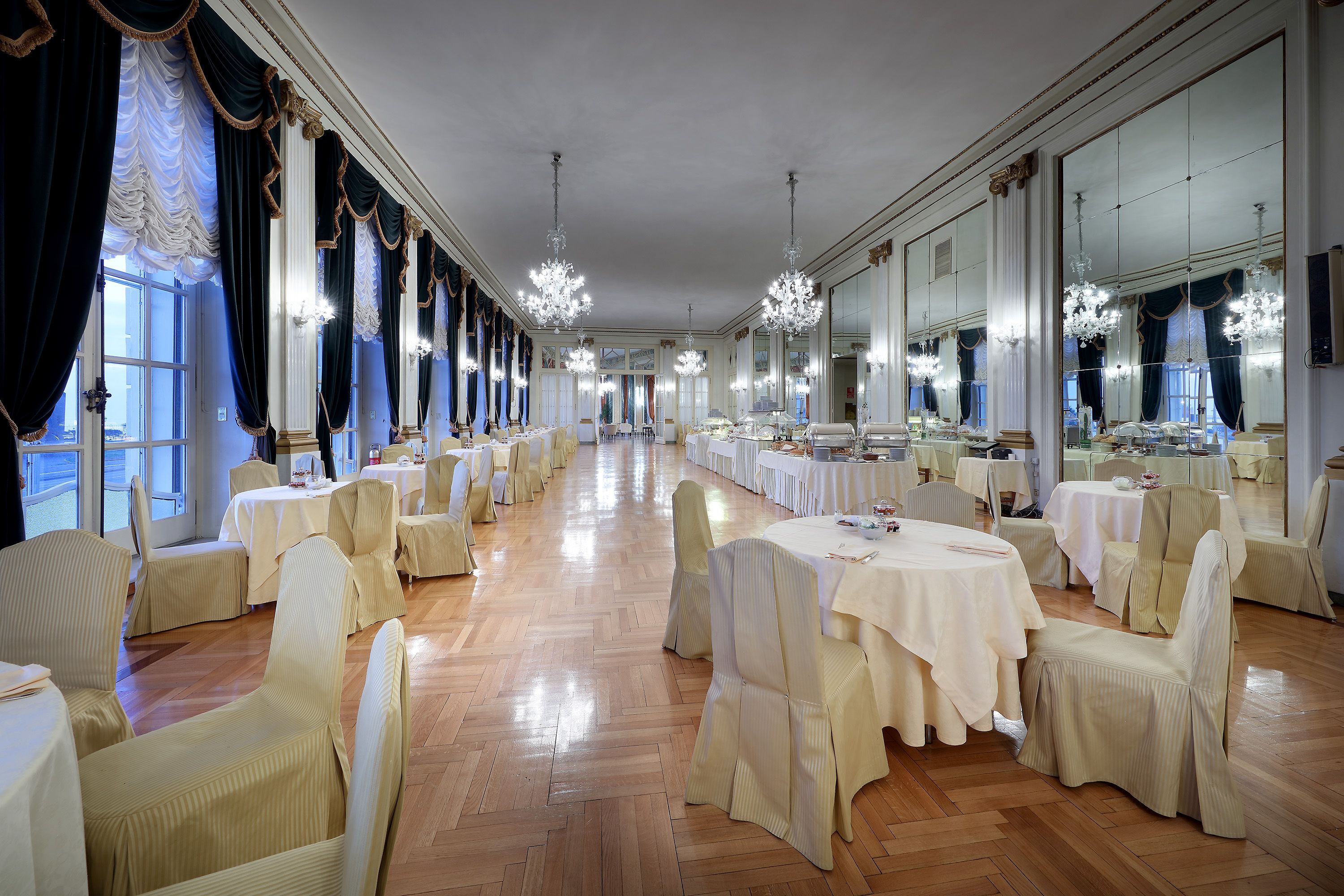 Une salle de banquet avec de longues tables, des chaises, des lustres et de grandes fenêtres.