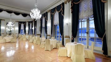 Una sala da banchetto con tavoli rotondi e sedie, ampie finestre con tende e lampadari.