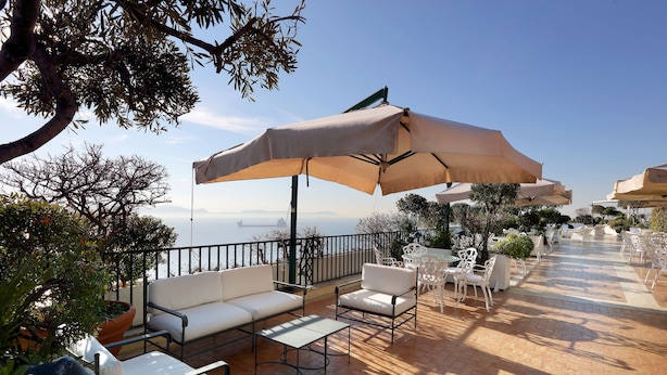 An outdoor patio with white furniture, beige umbrellas, and a view of the sea. - Eurostars Hotel Excelsior (Naples)