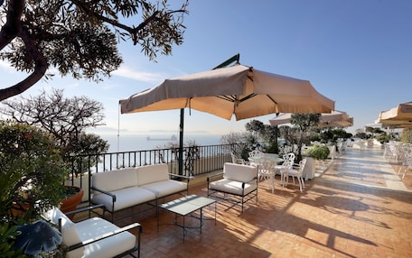 An outdoor patio with white furniture, beige umbrellas, and a view of the sea.