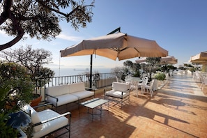 An outdoor patio with white furniture, beige umbrellas, and a view of the sea. - Eurostars Hotel Excelsior (Naples)