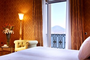 Hotelkamer met een bed, zithoek met een stoel, een tafel met bloevas, een lamp en uitzicht op een berg vanaf een balkon.