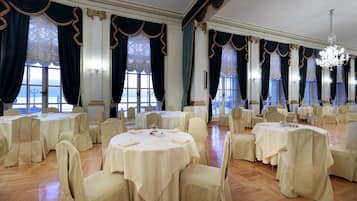 Un salón de banquetes con mesas y sillas redondas, ventanas amplias con cortinas y una araña.