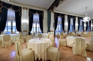 Un salón de banquetes con mesas y sillas redondas, ventanas amplias con cortinas y una araña.