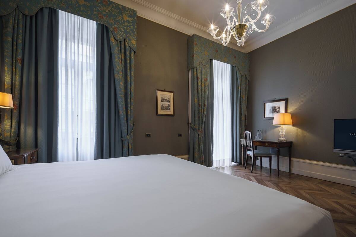 A hotel room with a large bed, a chandelier, a desk with a lamp, and a television.