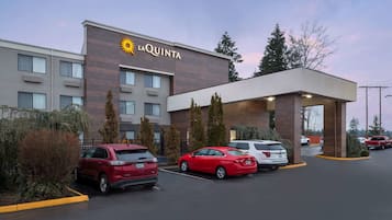 A La Quinta hotel with a modern design, featuring a prominent sun logo, a covered parking area, and several parked cars.