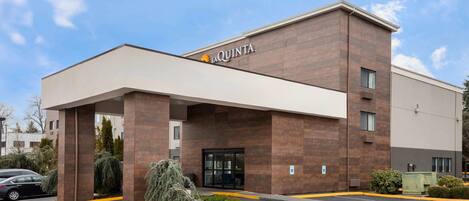 Un hotel moderno con un llamativo letrero de 'La Quinta', un acceso cubierto y un estacionamiento con espacios delimitados.