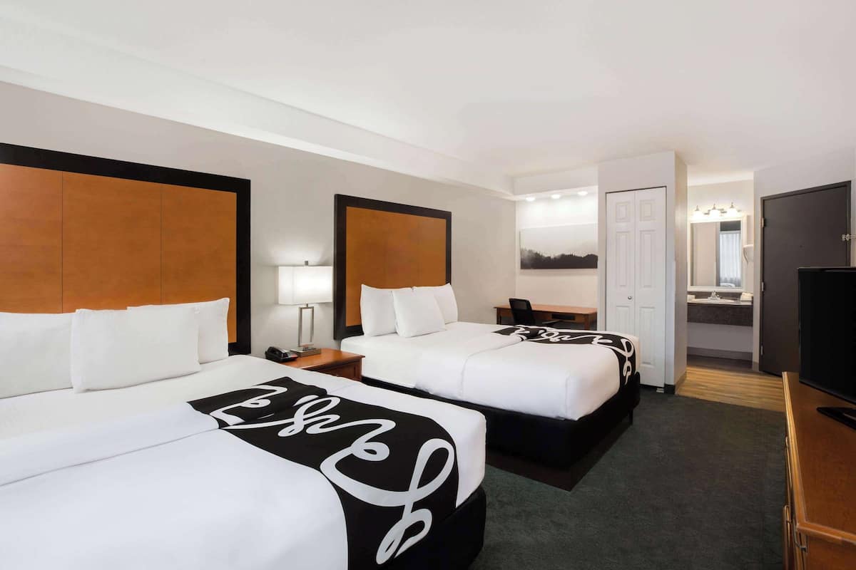 A hotel room with two beds, a desk, a TV, and a bathroom visible through an open door.