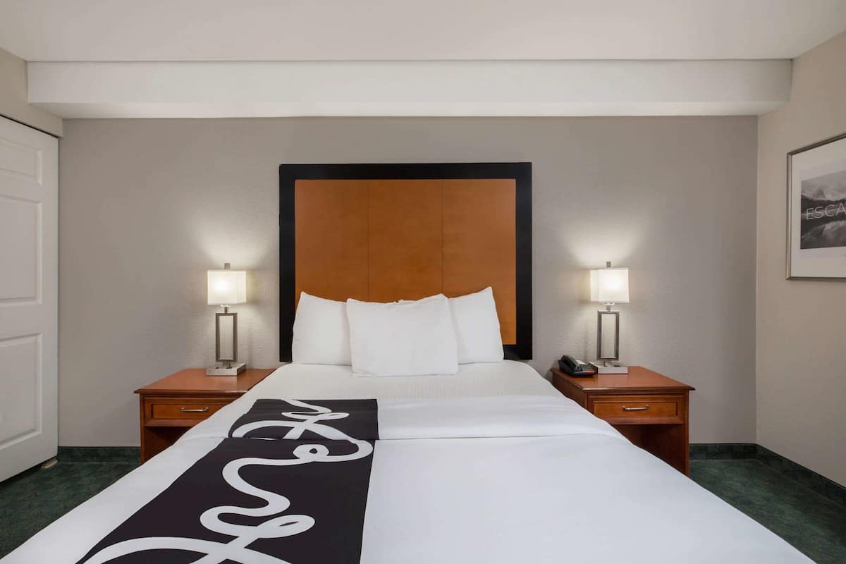 A hotel room with a large bed, two bedside tables with lamps, a wooden headboard, and a framed picture on the wall.