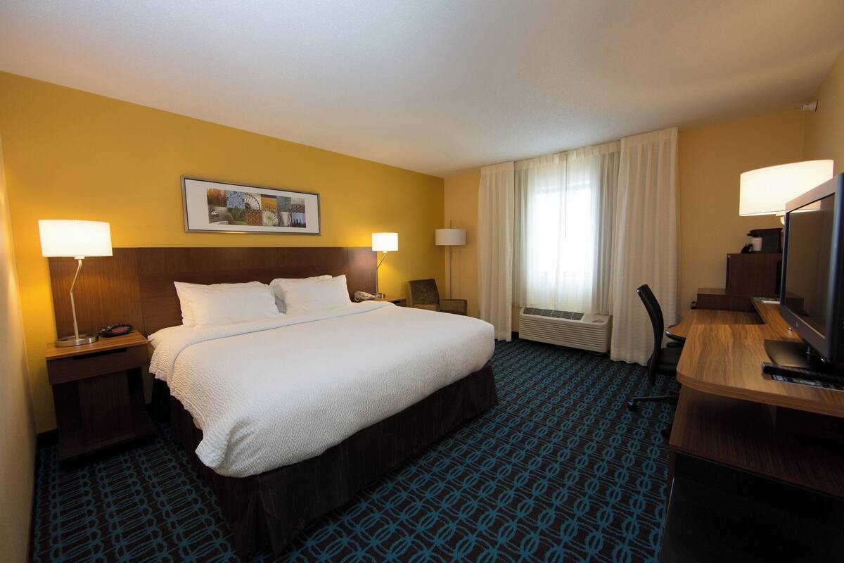 A hotel room with a large bed, two bedside tables with lamps, a desk with a computer and TV, and a window with curtains.