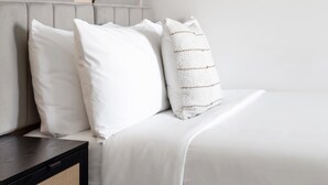 Ein ordentlich gemachtes Bett mit weißer Bettwäsche und Kissen, ein dunkler Nachttisch und ein gewebter Korb.