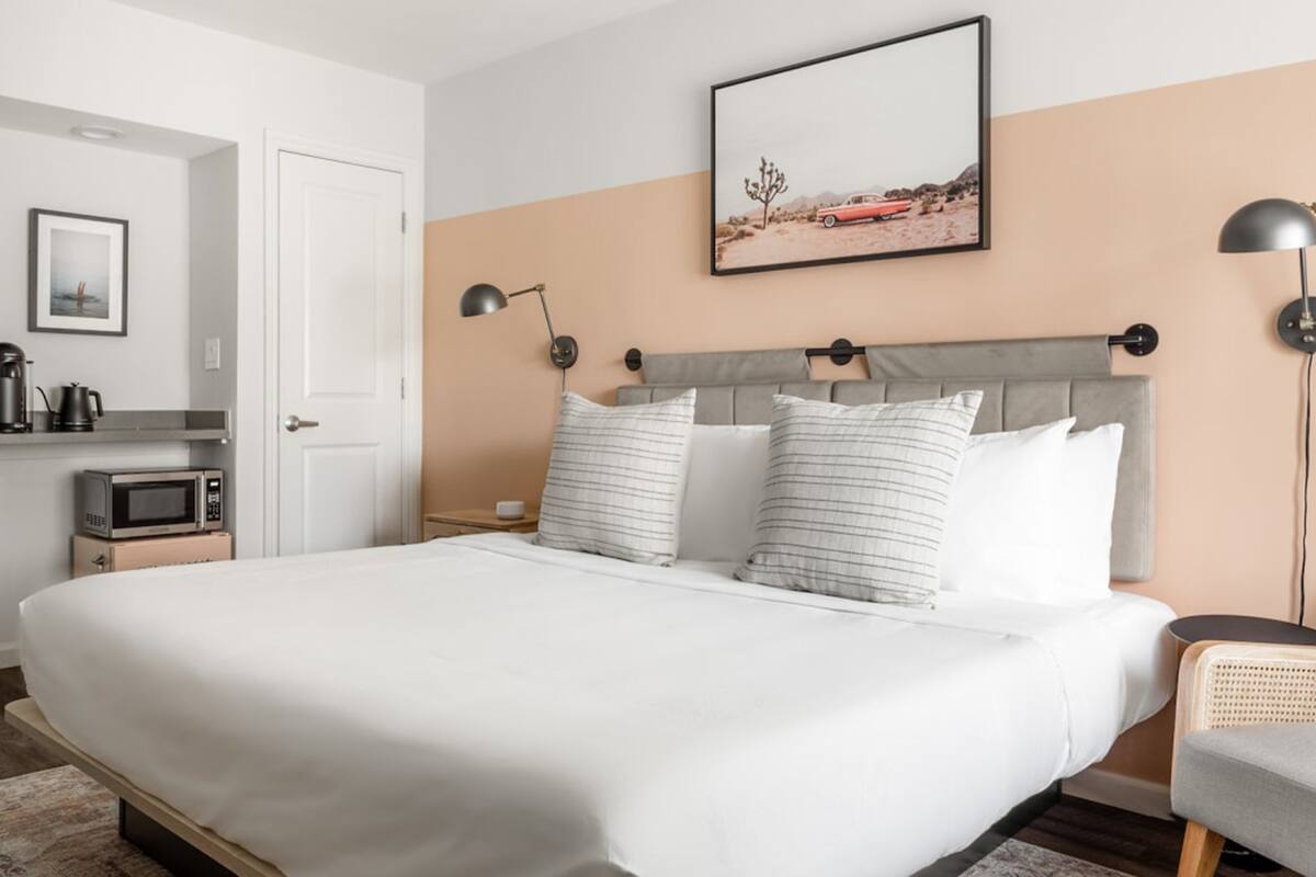 Una habitación de hotel con una cama grande, cabecera con varios almohadones, mesita de noche con lámpara, un cuadro en la pared y una zona de kitchenette con microondas y cafetera.