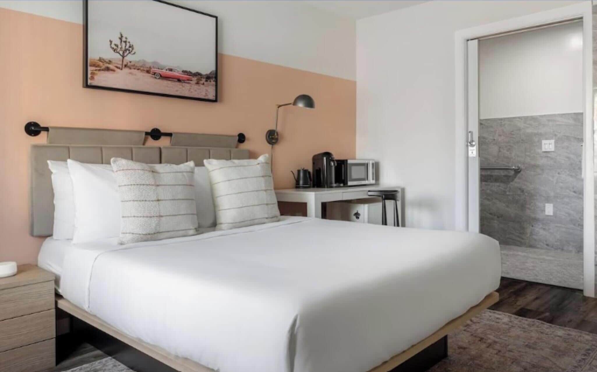 Un lit bien fait, avec du linge de lit blanc et des oreillers, une table de chevet avec une bouilloire et une machine à café, un tableau encadré au mur, et une salle de bain avec une douche et un porte-serviettes.