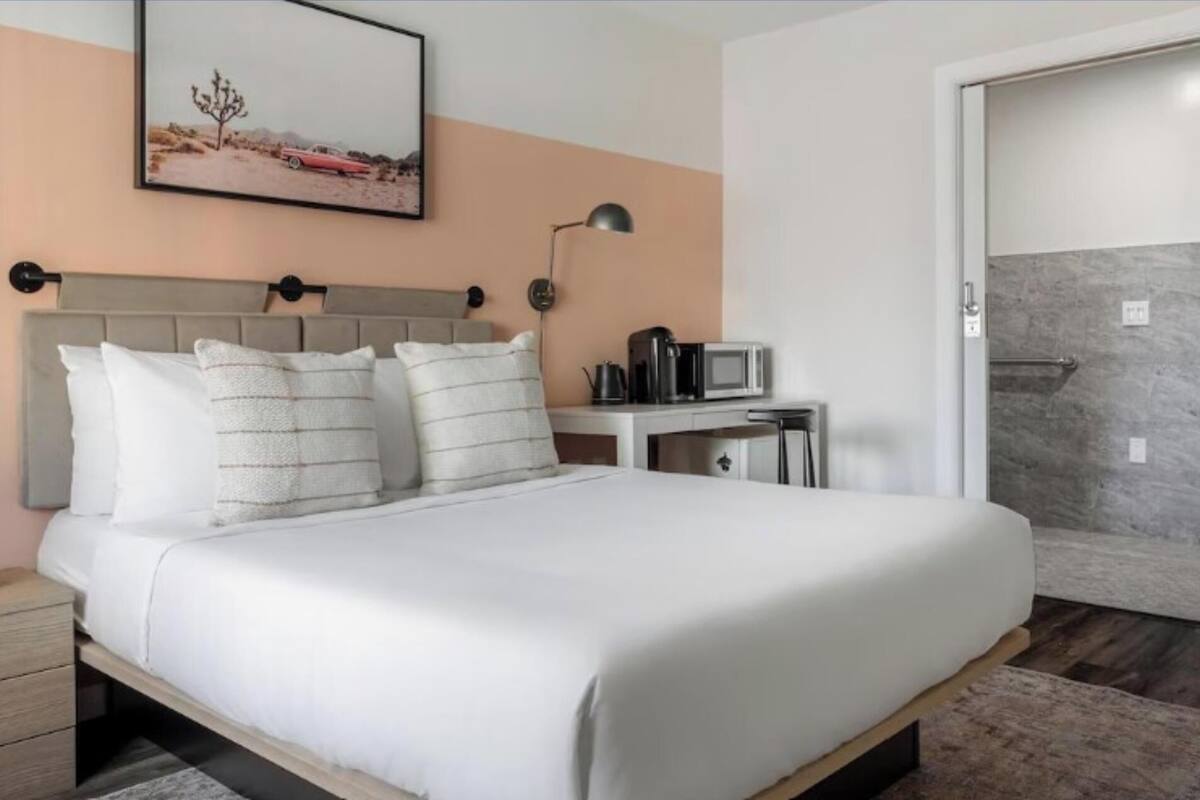 Una cama bien hecha con sábanas y almohadas blancas, una mesita de noche con hervidor y cafetera, un cuadro en la pared y un baño con ducha y toallero.