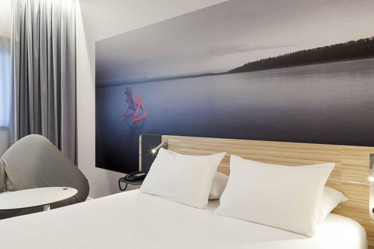 Habitación de hotel con cama, una silla, una mesita, teléfono y un cuadro de un lago en la pared.