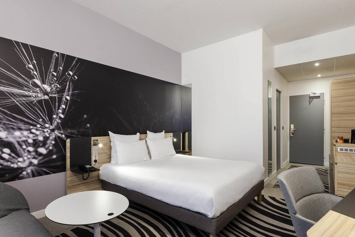Una habitación de hotel moderna con una cama grande, una mesita redonda pequeña, un sofá gris y una pieza de arte original en la pared.