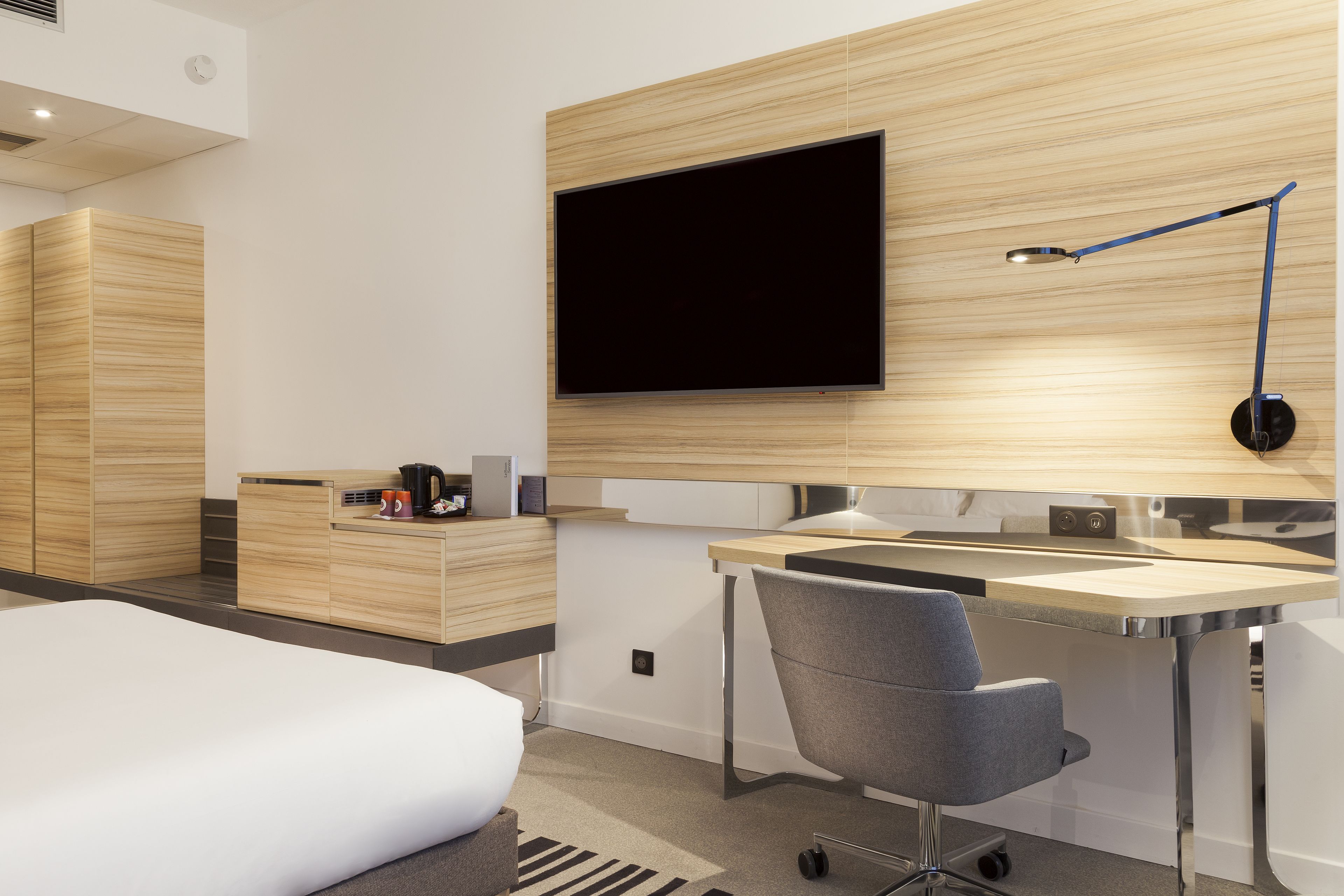 A modern hotel room with a bed, a desk with a chair, a flat-screen TV mounted on the wall, and a coffee maker on the side table.
