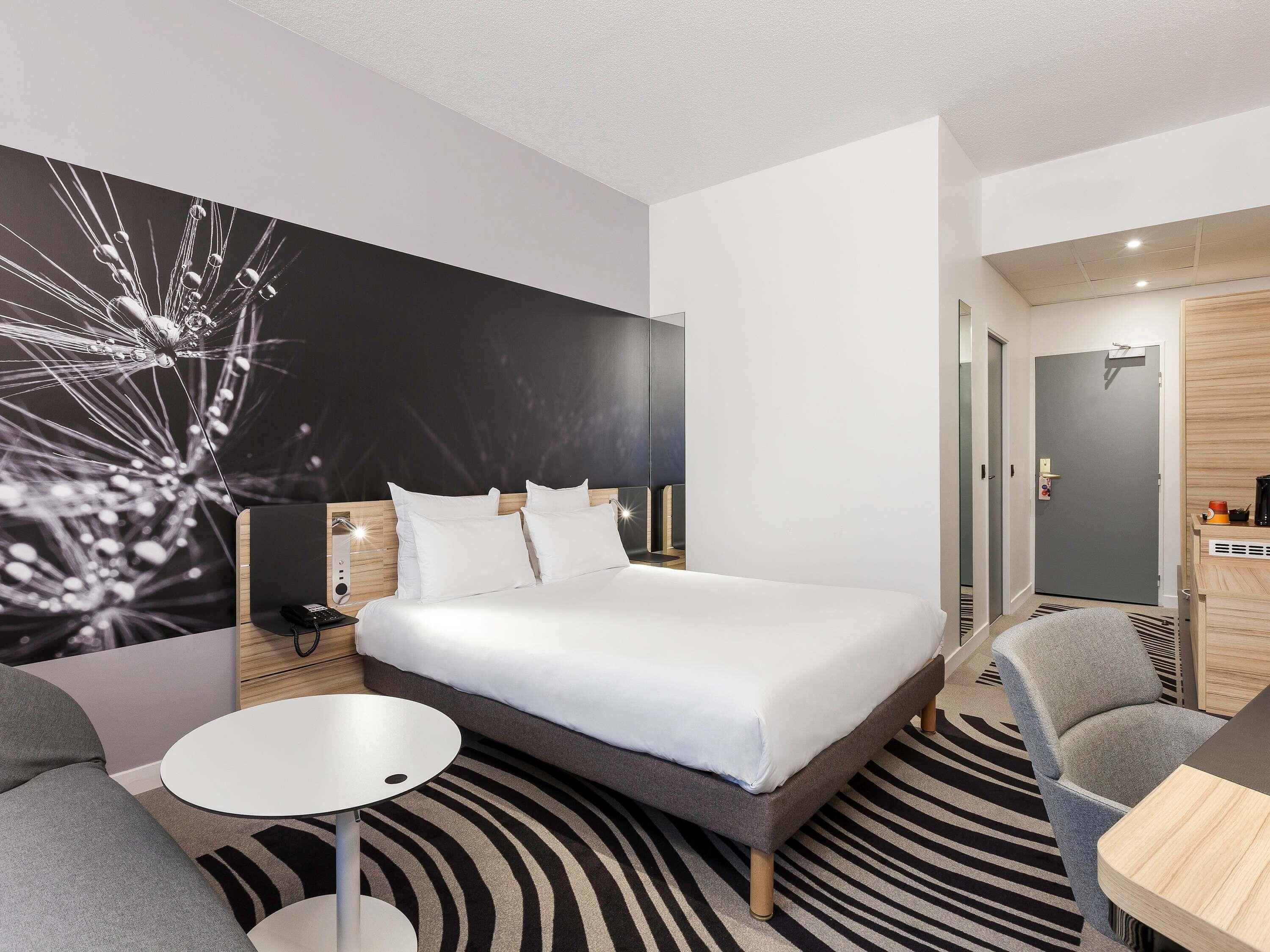 A modern hotel room with a large bed, a small round table, a grey sofa, and a zebra-patterned rug.