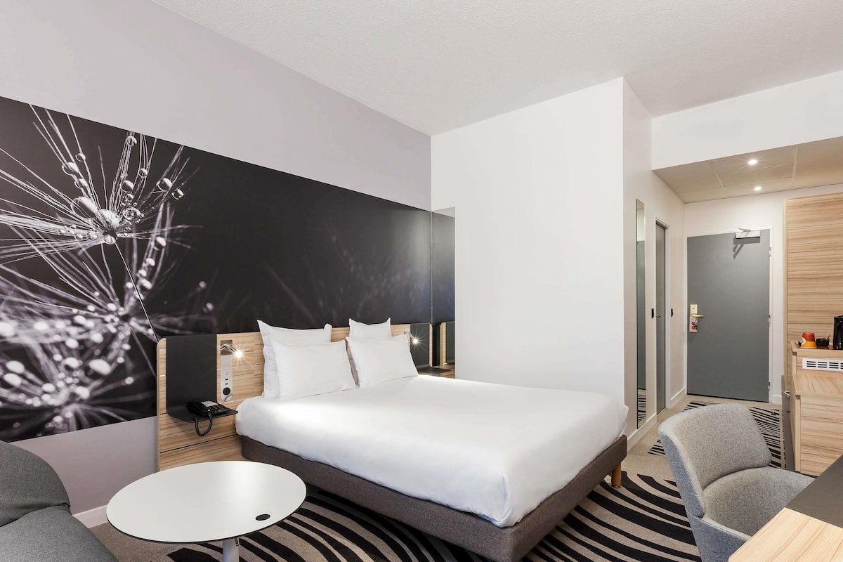 Una habitación de hotel moderna con una cama grande, una mesita redonda pequeña, un sofá gris y una alfombra con estampado de cebra.