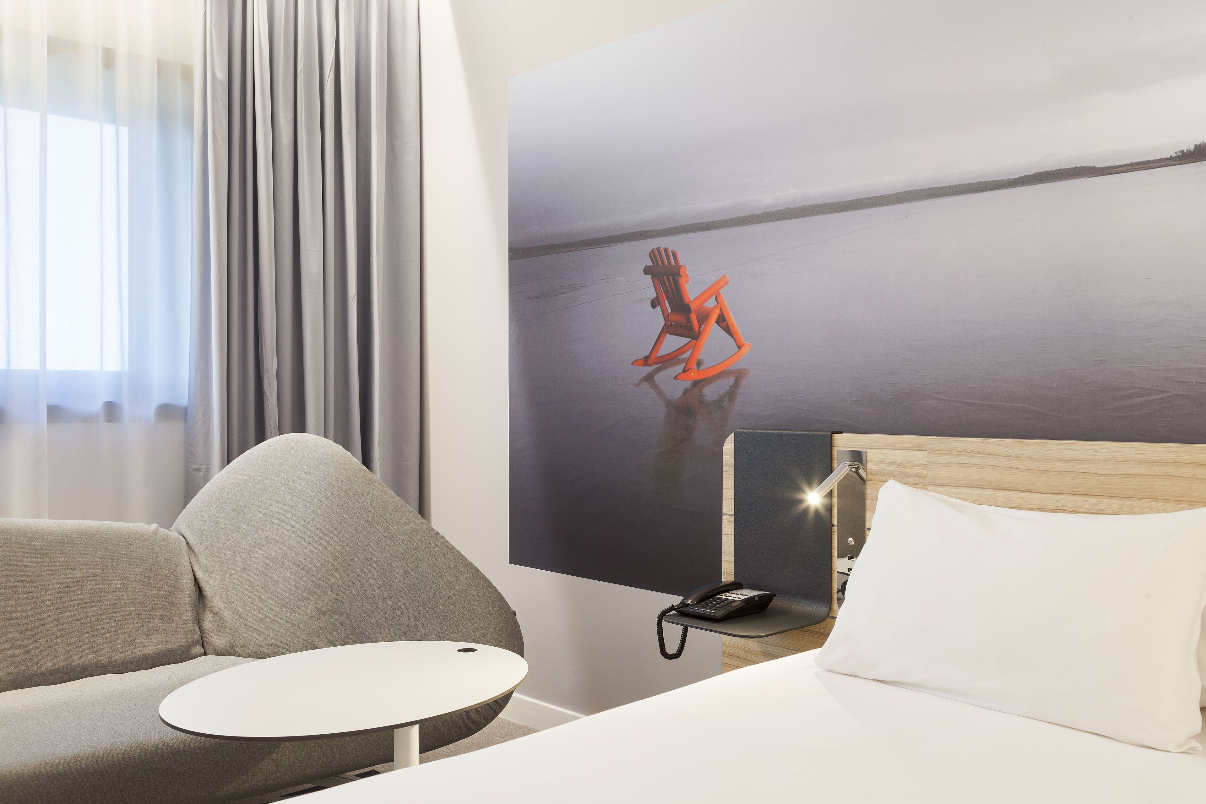 Una camera d'albergo moderna con un letto, una poltrona, un piccolo tavolino rotondo, un telefono e un quadro raffigurante una sedia su una spiaggia.