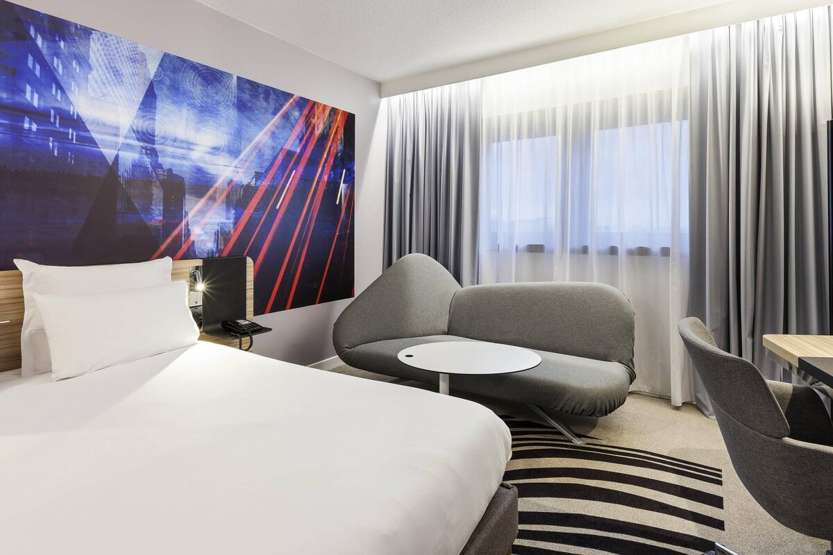 Habitación de hotel con cama, sofá, una mesita y un cuadro abstracto grande en la pared.