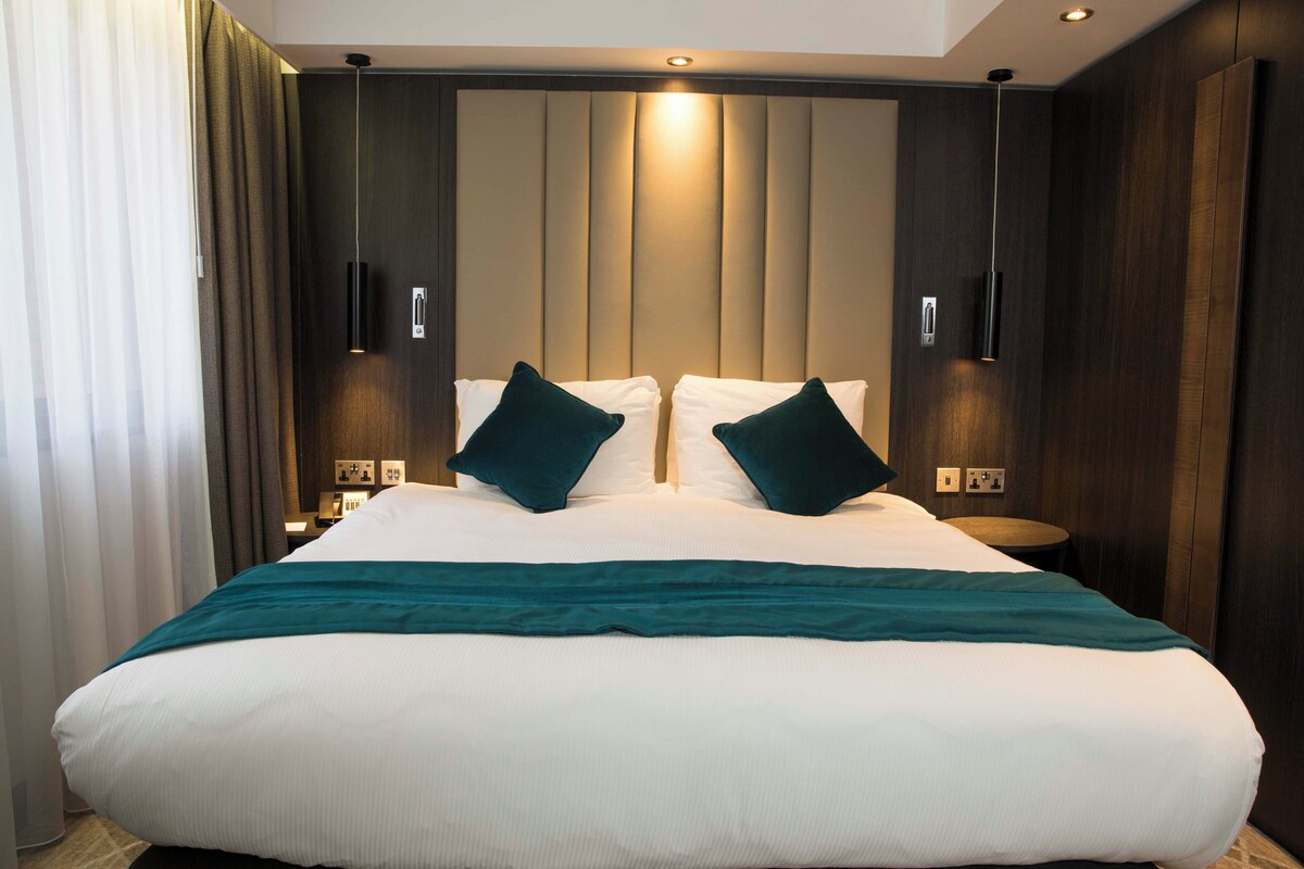 A hotel room with a large bed, two bedside tables, a headboard with vertical panels, and wall-mounted lighting.