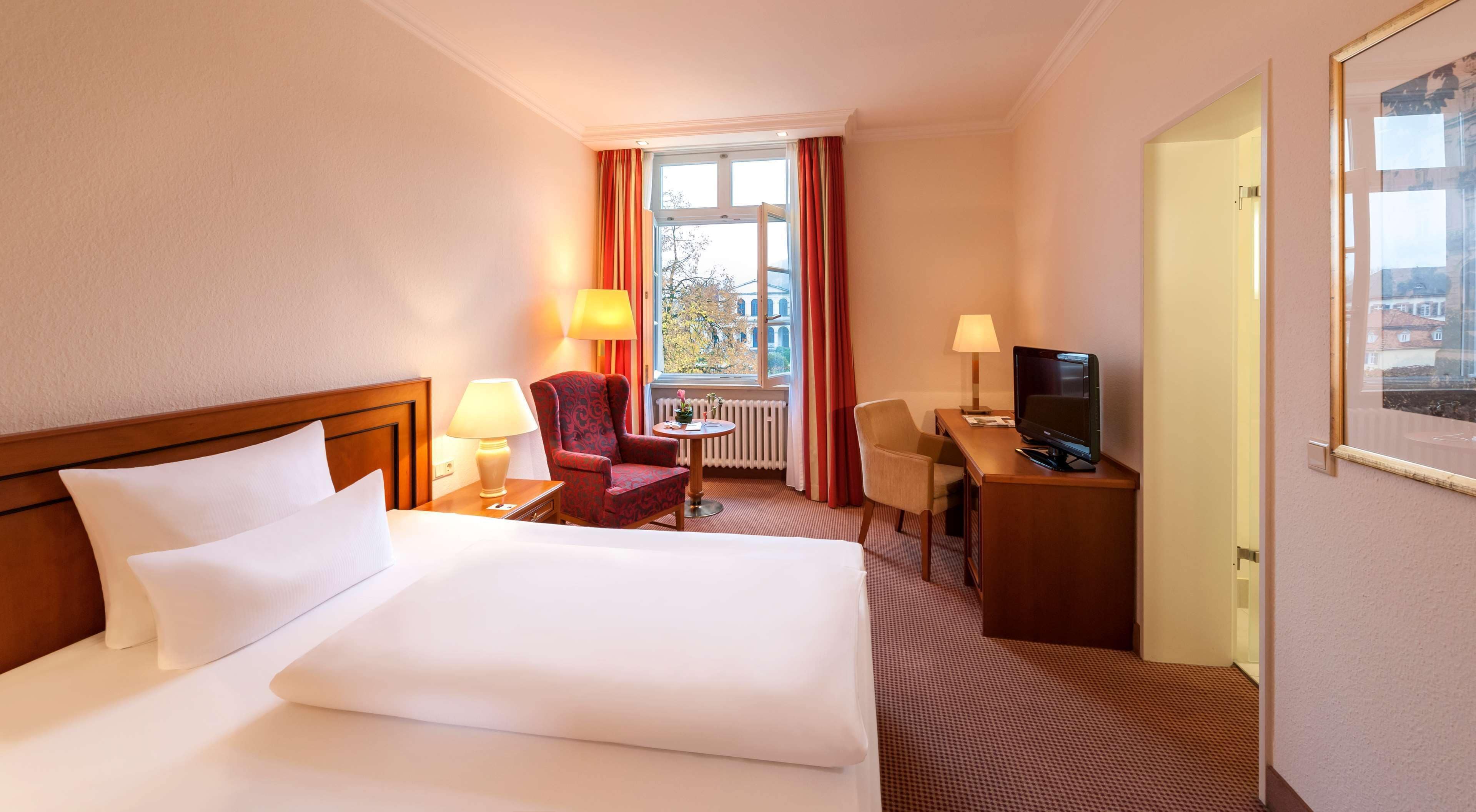 Une chambre d’hôtel avec un grand lit, deux fauteuils, un bureau avec une télévision et une fenêtre avec des rideaux.